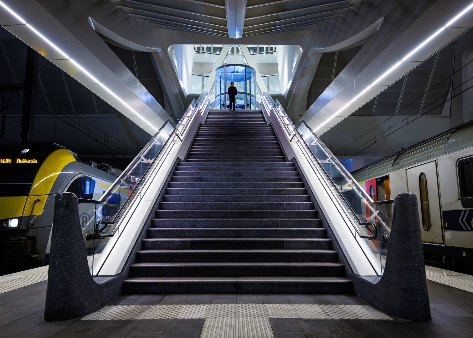 Look-up symetrical night shot with a wide-angle lens of a young man standing at the top of stairs at the Mons train station designed by architect Calatrava. The man is about to go down the deep stairs leading to a platform where trains are waiting on each side. A blue-lit glass elevator can be seen behind his back.  The stairs are protected by a majestic white modern ceiling structure.