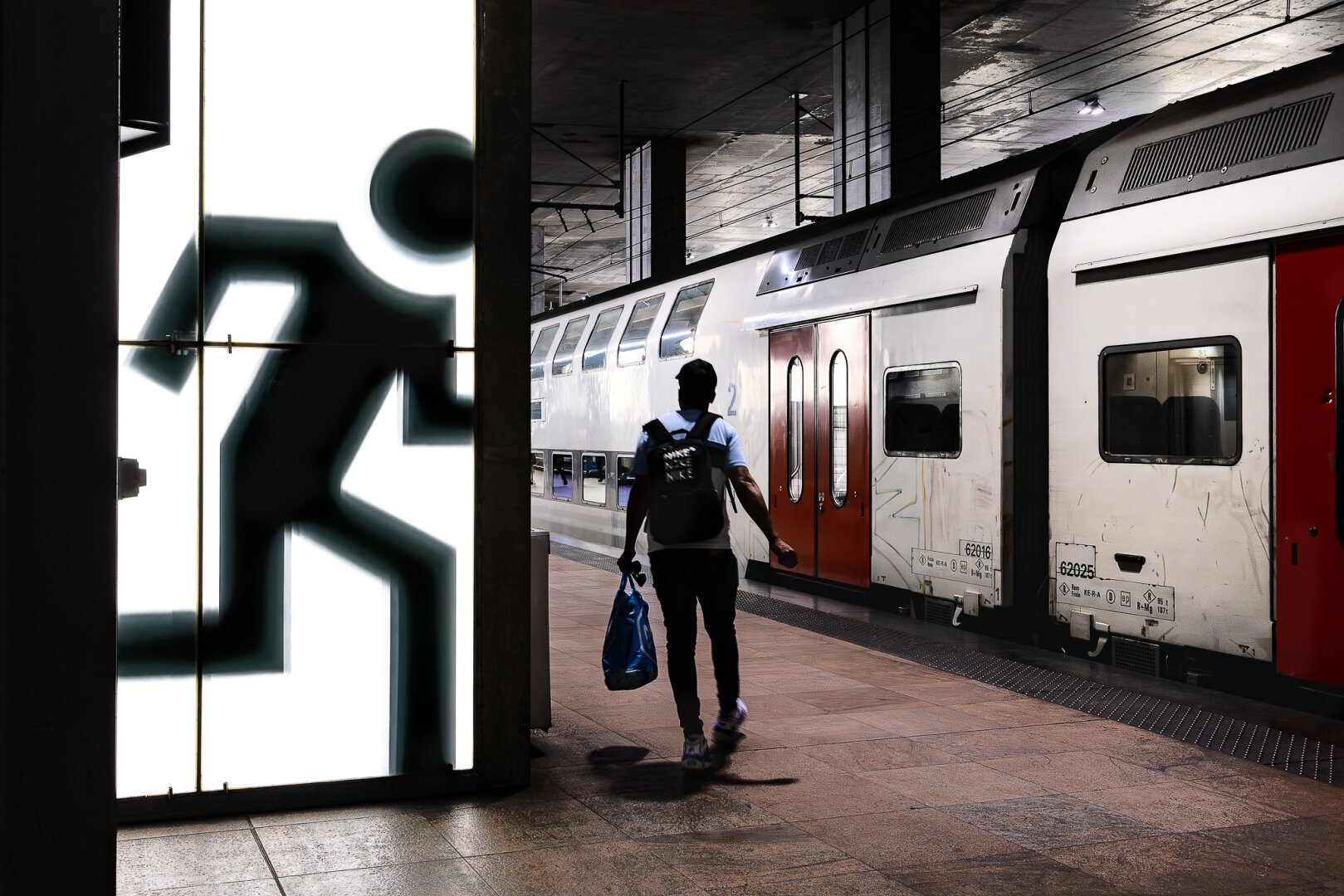 At the Antwerp train station in Belgium, on one of the underground platforms, a passenger, in the center of the image and seen from his back and slightly in the shadow, is turning a corner on the left side after he passed a white lit panel with a black pictogramme silhouette of a running human character bigger than him. The passenger is speeding up to catch his train which is standing on the platform on the right of the image.  The train is predominantly white with some black patterns and fits well with the visual effects of the two black (human and pictogramme) silhouettes, making the image very graphic. A red door at he back of the train in the background, the pastel warm color of the tiles on the floor and patches of blue on parts of the passenger that are not in the shadow add color touches to this image where the black on white are predominant.