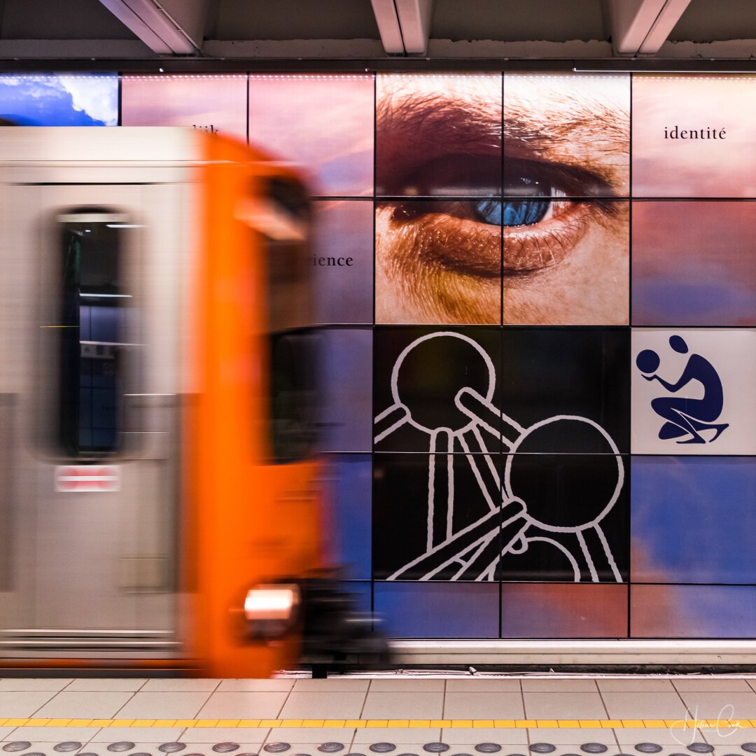 Slow motion shot of an orange metro train enters the Heizel metro station in Brussels, passing in front of awall decorated with a big size photo of a human blue eye, and other artworks.