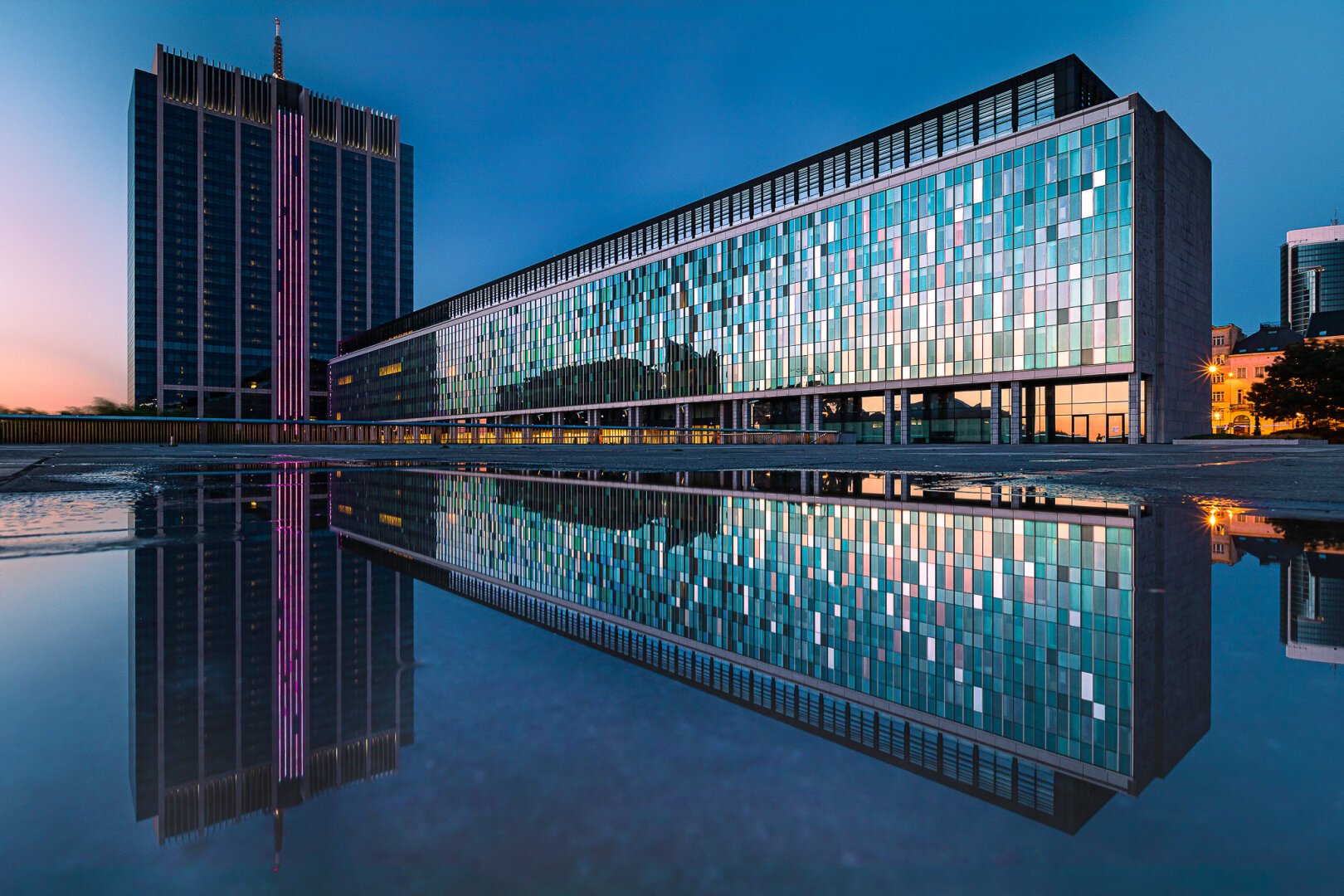 Reflection shot in a puddle of an administrative tower in Brussels and its rectangular anne. The picture was taken at the end of sunset when the sun still makes an orange glow on the windows, while the sky is turning dark turquoise. The building with its reflexion in the water evocates a giant spaceship from science fiction movies.