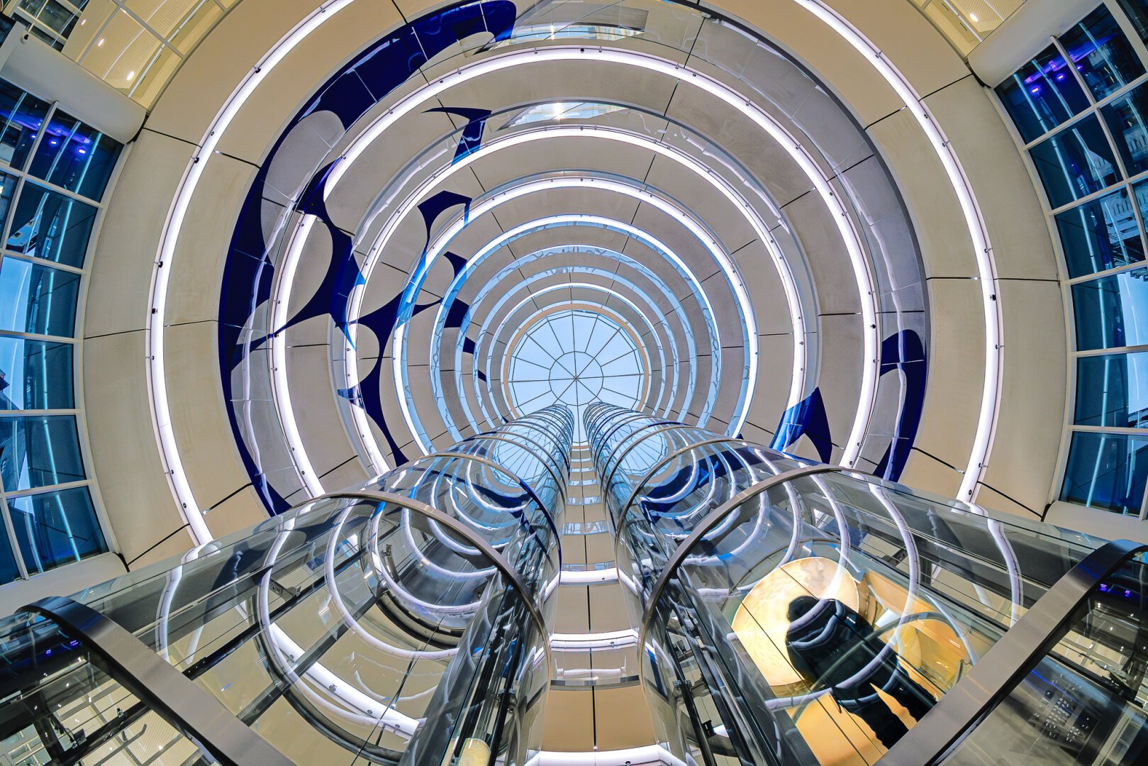 Look-up wide angle shot at blue hour of a rond atrium with a glass ceiling in a modern building and the silhouette of a person going up in a yellow-lit glass elevator, evoking a space journey.