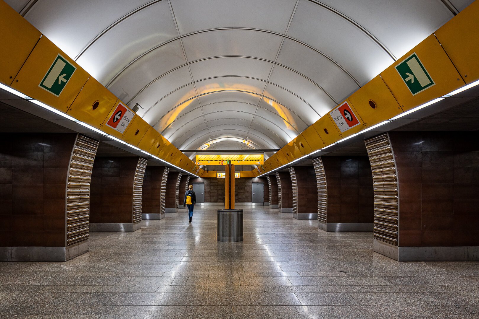 Symetrical wide angle shot of main hall leading to the platforms at the Jinonice metro station in Prague, featuring a modern architecture with a curved white shiny ceiling, yellow/orange panels indicating the directions and brown pillars. The silhouette of a lady with a yellow bag matching the colors of the station can be seen going towards the end of the hall.