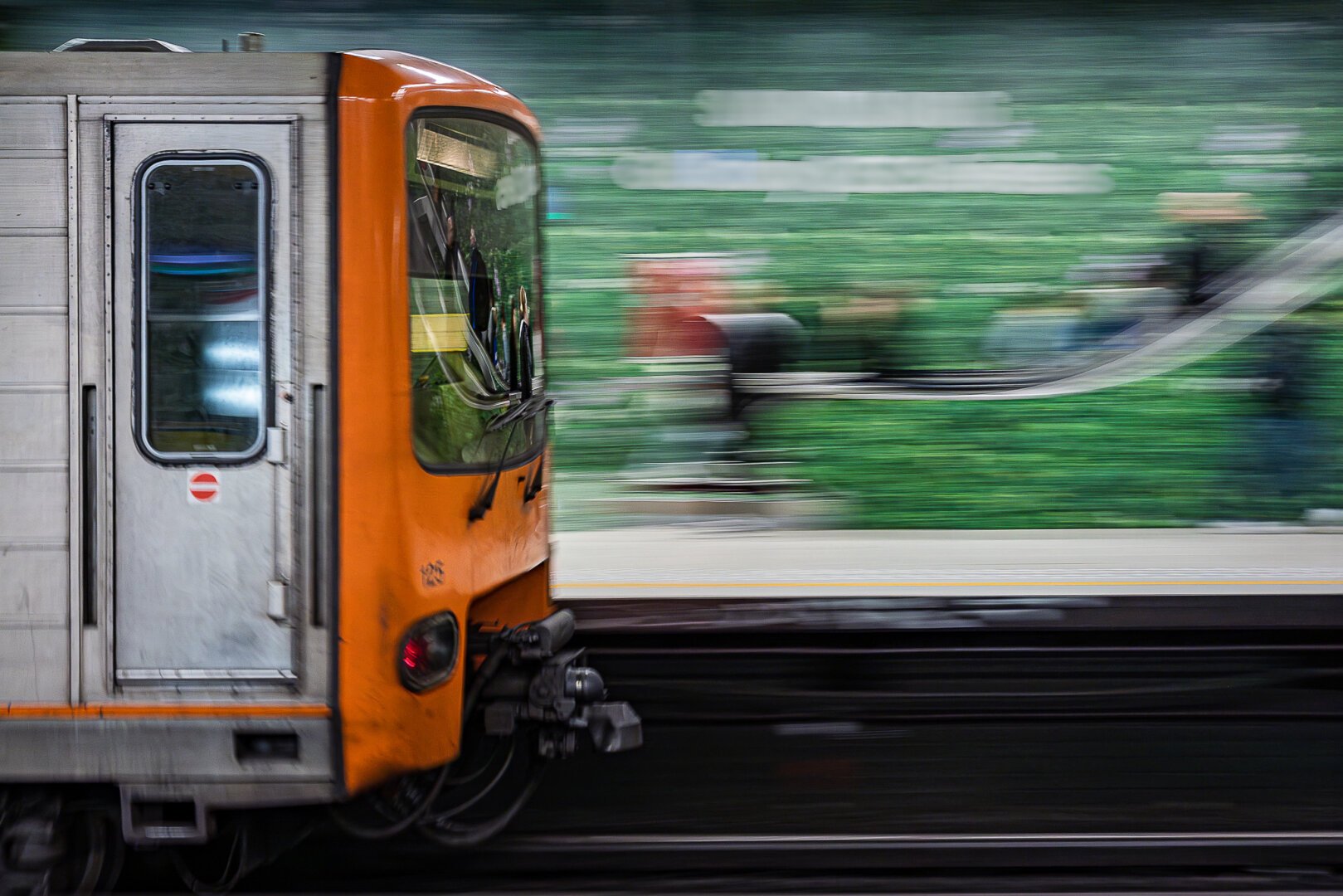 Panning shot at a slow shutter speed of an orange and grey head of a train entering a metro station in Brussels whose walls had been temporarily decorated with gigantic stickers of green grass for a commercial campaign. The train looks fairly sharp and people ascending an escalator are visible on the reflection in the front window whereas the background composed of the green glass and the actual elevator are brushed away in an aesthetic fashion. This shot was taken at the Louise metro station.