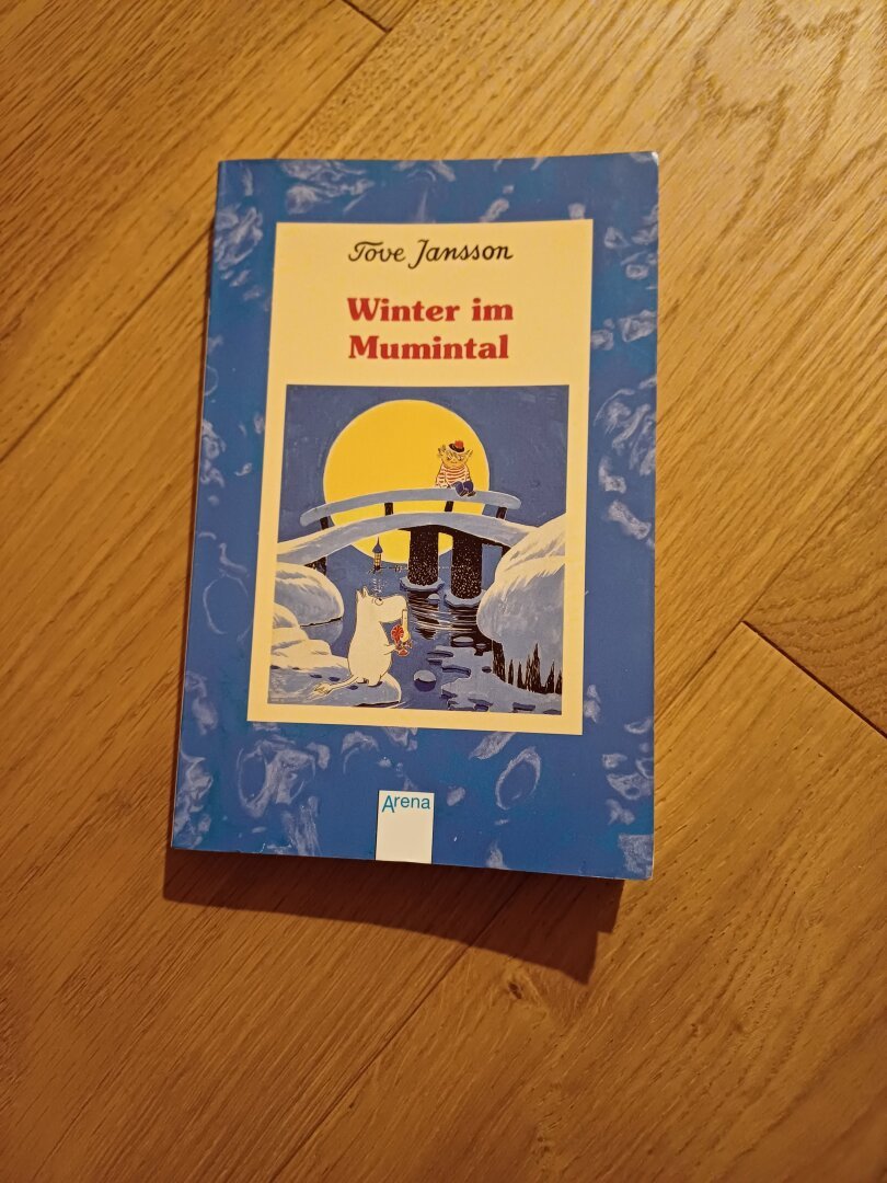 Das Bild zeigt das Buch Winter im Mumintal von Tove Jansson auf dem Cover ist eine verschneite Brücke am Meeresufer zu sehen. Auf der Brücke sitzt eine Figur mit rotweiß gestreiftem Pulli und roter Mütze. Vor der Brücke steht ein Mumintroll mit weißem Fell, großer Schnauze und Schwänchen mit Bausch. Im Hintergrund sieht man den Vollmond