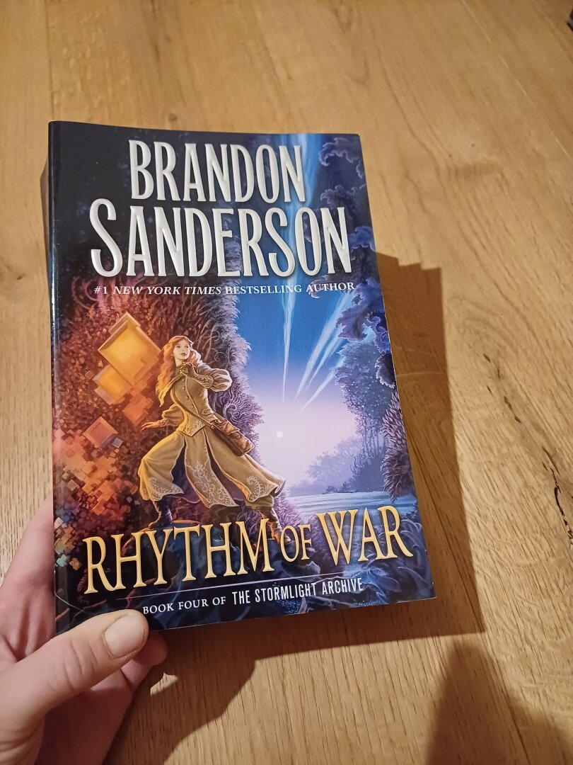 The picture shows the book Rhythm of war by Brandon Sanderson. The cover shows a women with a light complexion and red hair, wearing beige clothing and a brown satchel. The background is blue with plants on one side and yellowish with graphic patterns on the other.