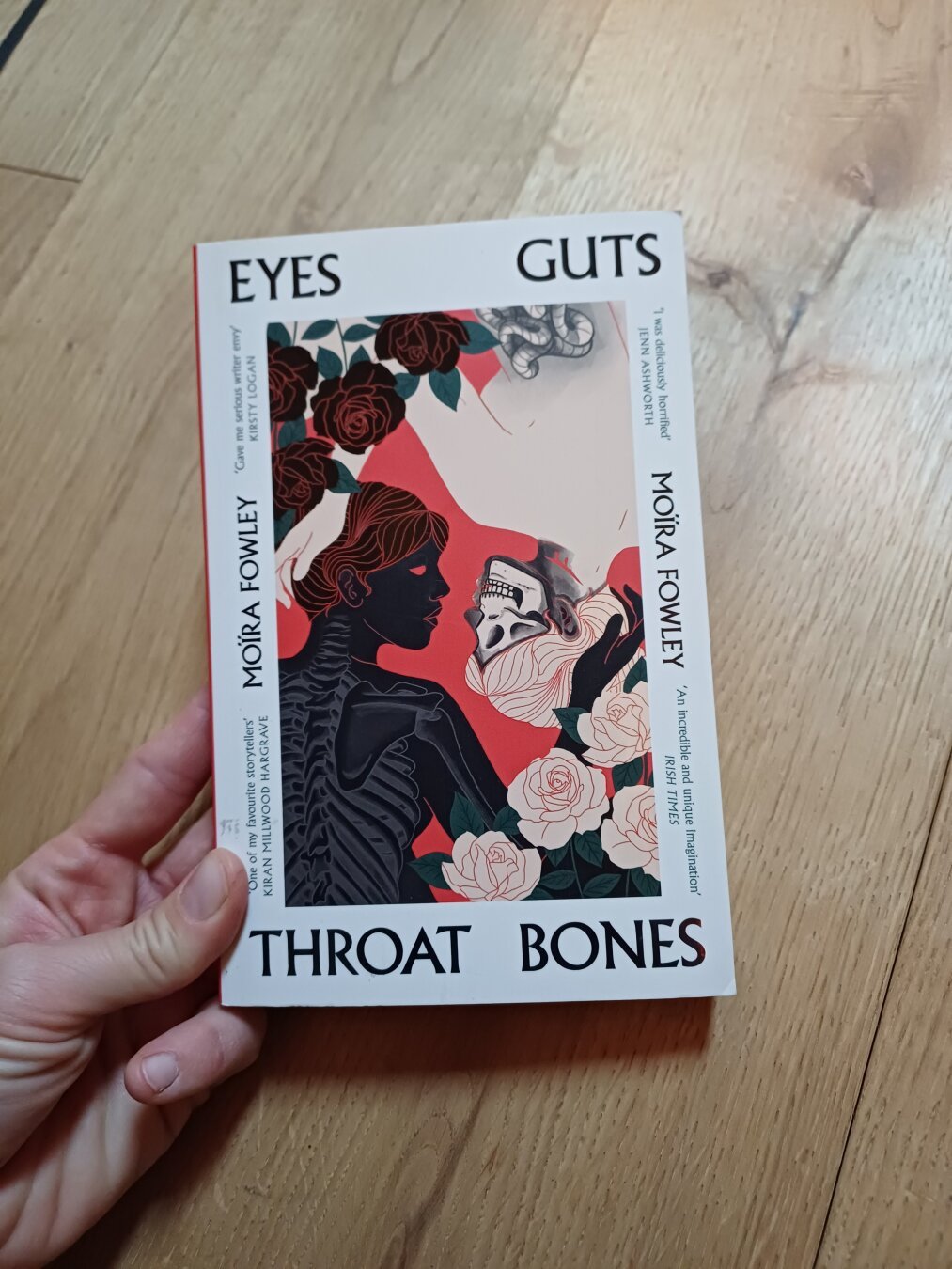 The picture shows the book eyes guts throats bones by Moïra Fowley. On the cover we see drawings of two upperbody silhouettes of women one in white with the head in the style of a skull, the other black with contours and a torso showing the bones. They are placed upside down to each other similar to many carddeck designs. They are framed by white and dark red roses. The background is red. 
The cover is framed in white and the title is written on this frame in black.
