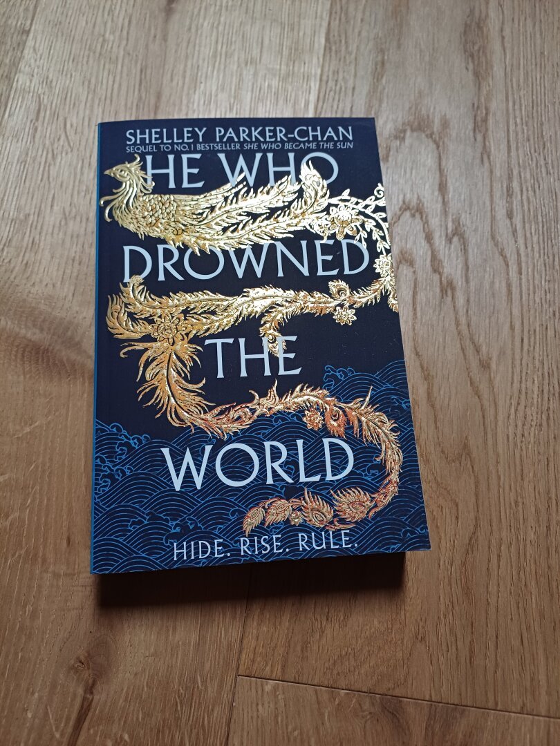The picture shows the book he who drowned the world by Shelley Parker-Chan. The cover is dark blue and shows a winding stylized golden bird with long and intricately patterned tailfeathers. The title is in white