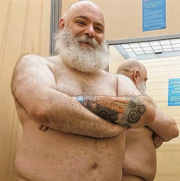 Me, a big-bearded caucasian-presenting Santa-Biker-looking cis-dude, nekkid (but only showing from the waist-up, for now) in a changing room, with my arms crossed & a big smile on my face.