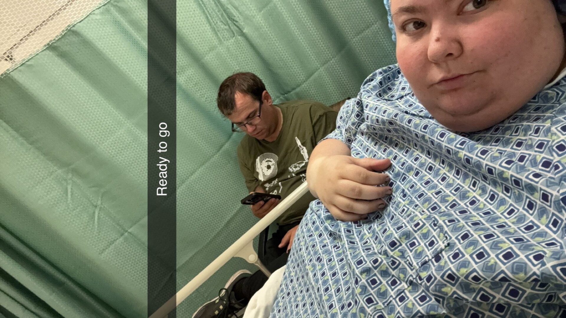 Woman in hospital gown in hospital bed with man next to her in chair playing on phone. Text on picture says "ready to go" (at 310lbs)