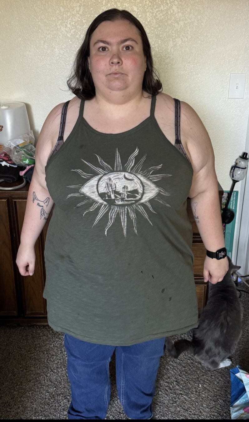 Woman wearing green tank top with white design of eye with dessert scene in eye ball and turned into sun standing in front of dresser. (At 234lbs)