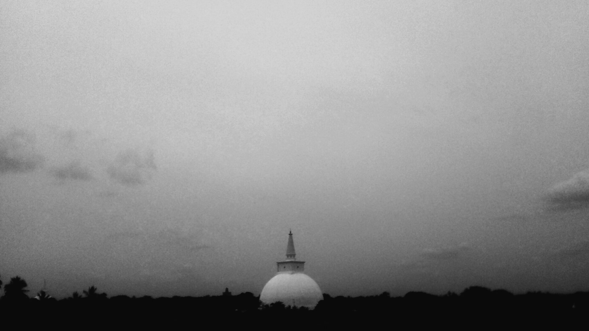 A black and white photo of a stupa rising from a dark tree line under a vast, quiet sky. The serene and minimalist composition evokes a sense of stillness, spirituality, and timeless presence.