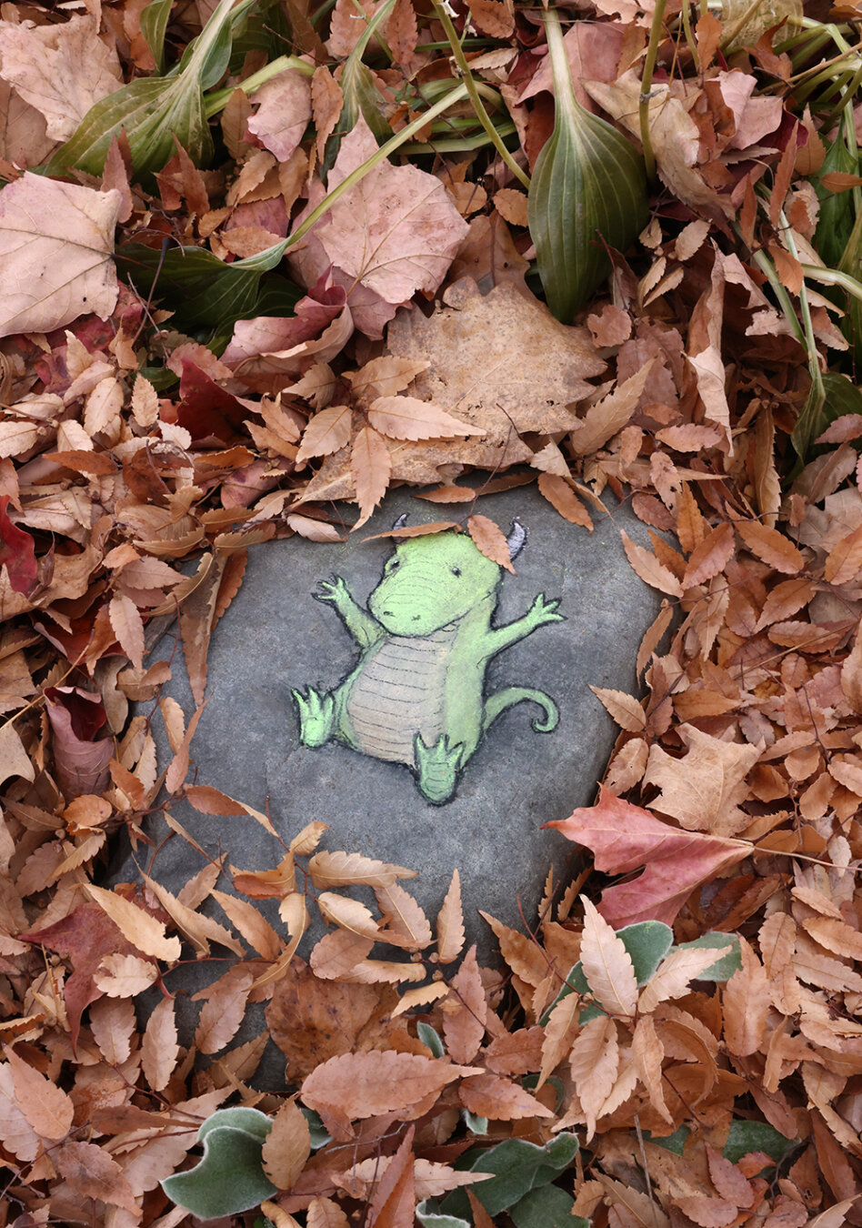 chalk art drawing of a small dinosaur sitting on a rock surrounded by reddish fallen leaves, two of which have fallen on top of his head.