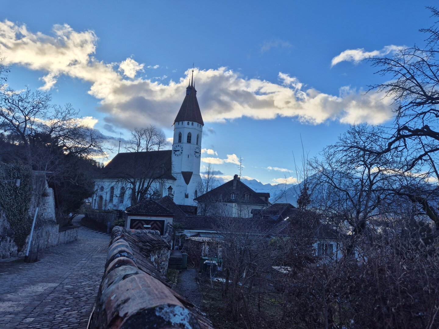 Im Zentrum des Bildes steht eine Kirche mit einem hohen, spitzen Turm, der sich gegen den blauen Himmel mit weißen Wolken abhebt. Die Kirche ist weiß gestrichen und hat ein dunkles Dach. Um die Kirche herum gruppieren sich weitere Gebäude, die typisch für die Architektur dieser Region sind. 

Im Vordergrund des Bildes ist ein gepflasterter Weg zu sehen, der sich entlang einer Mauer schlängelt. Die Mauer ist mit roten Ziegeln bedeckt und bietet einen schönen Kontrast zum weißen Gebäude der Kirche. Links und rechts des Weges befinden sich Bäume, die zum Teil kahl sind, was auf eine kalte Jahreszeit hindeutet. Im Hintergrund ist der Niesen zu erkennen, von einer leichten Schneedecke bedeckt.