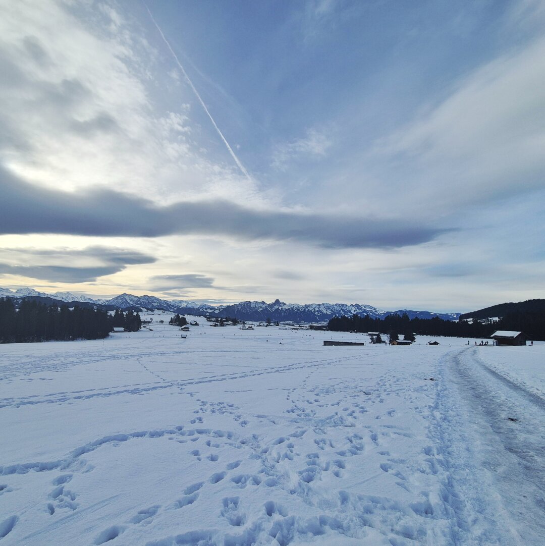 Das Bild zeigt eine weite, verschneite Landschaft mit einer Himmelsansicht.

Eine unberührte Schneedecke bedeckt den Boden und erstreckt sich bis zum Horizont. 
Zahlreiche Fußspuren unterbrechen die weiße Fläche und führen in verschiedene Richtungen. 
Am rechten Bildrand ist ein schneebedeckter Weg erkennbar.

In der Ferne ist eine Bergkette erkennbar.

Der Himmel ist von hellblauen Farbtönen dominiert, mit einigen weißen Wolken, die sich darüber verteilen. Ein Kondensstreifen eines Flugzeugs zieht sich diagonal über den Himmel.