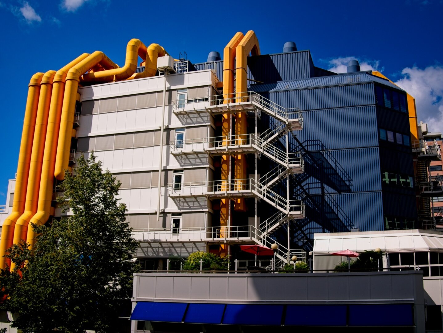Multistory post modern building with large yellow tubes on the outside left and middle sections snaking over the building and a large blue box on the right side of the building with white in the center.
Outside staircase in the middle cast a nice sharp shadow over the right side and a bright blue sunlit sky in the background.