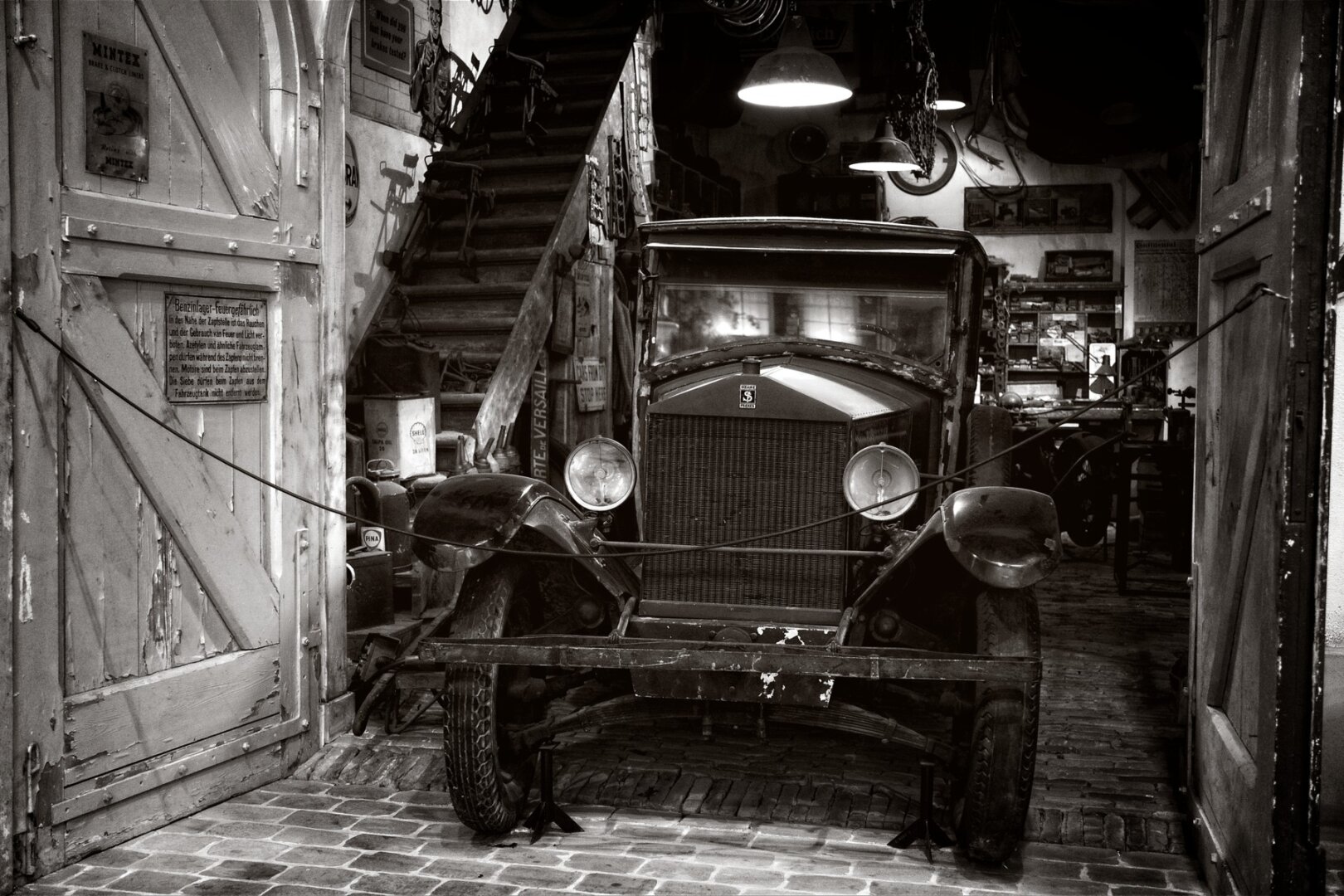 Black and white photo of a on old barn workshop. Two doors with flaky white paint and a german sign are open, revealing the very old Rolls Royce car in the middle. Surrounding it are lots of tools and paint cans with a staircase in the back. Dimly lit by two overhead industrial lights.
Everything is just a little bit dusty and weathered.