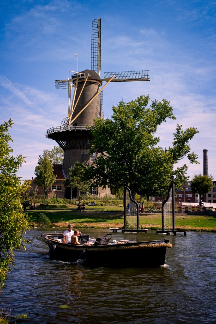 De Valk windmill (stone, taperd tower with a wooden swivel top) shot from behind on a sunny day, surrounded by some trees and a park.
A small, black, open top motor boat sails past in the foreground.