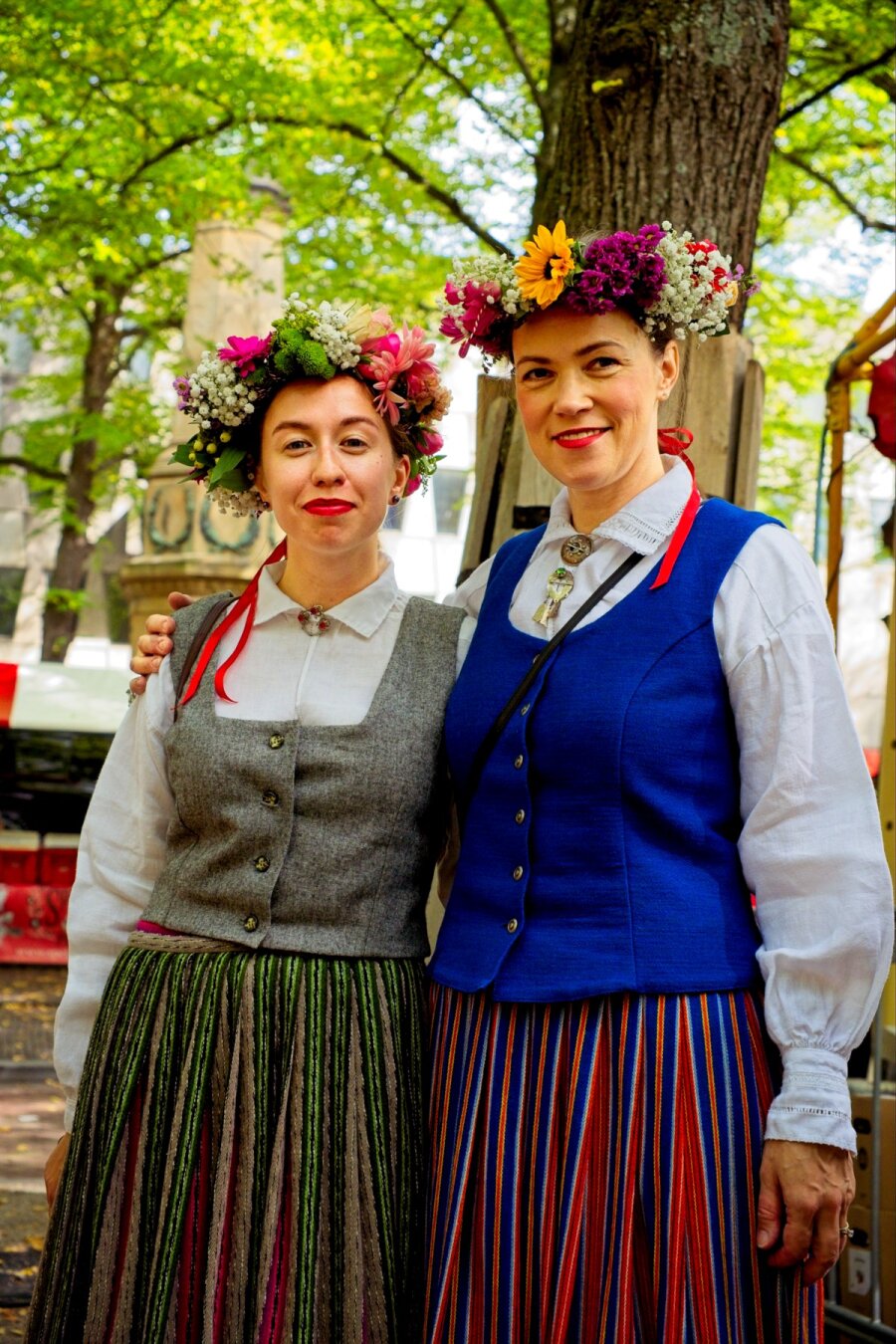 Two women wearing flower crowns and tradional cloths. White blouses with a blue coat and a red blue striped dress on the right. White blouse with a gray coat and green red striped dress on the left.
A bright forrest of trees is seen in the background.