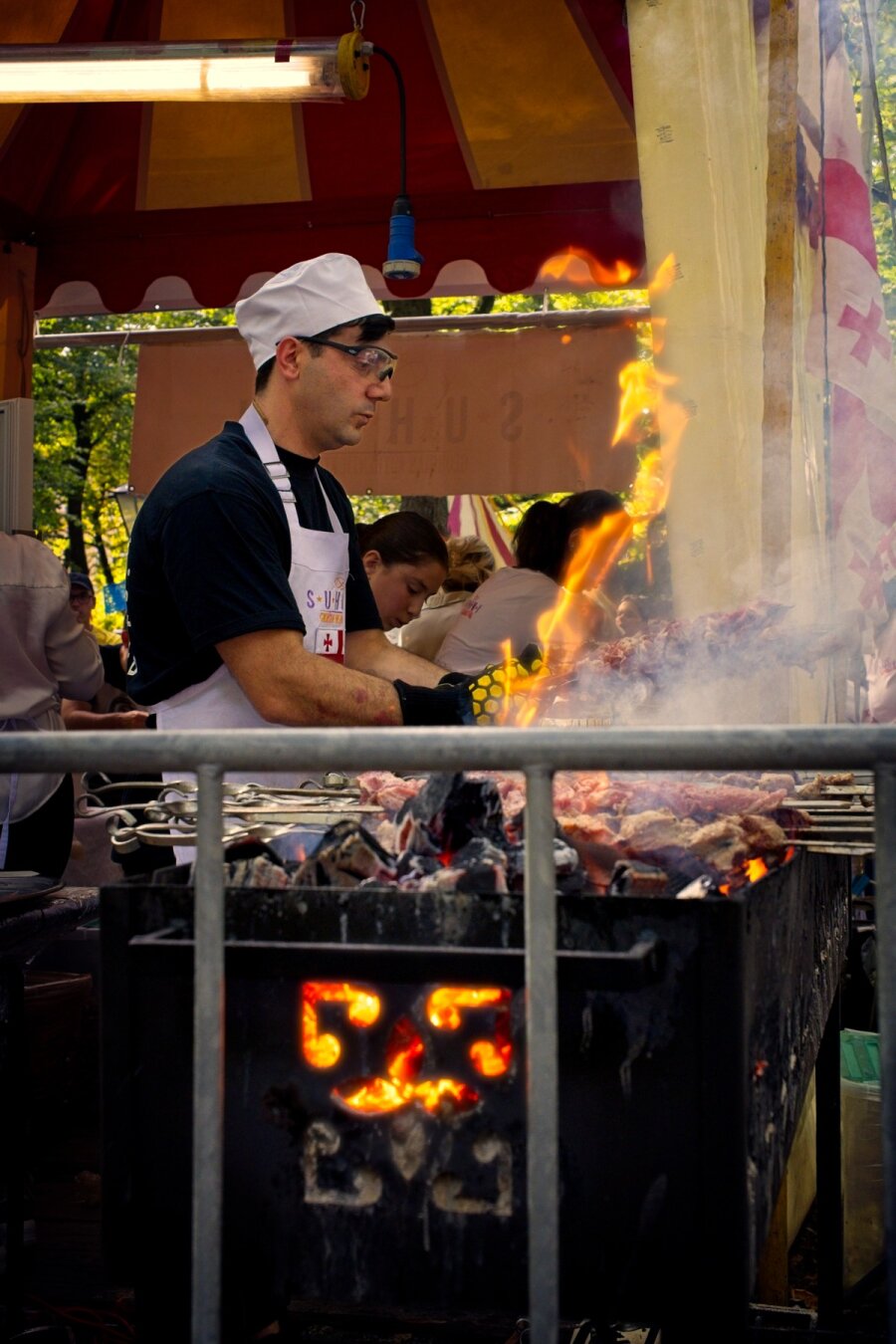 A man in cooks cloths standing behind a blazing BBQ holding two meat spikes. The BBQ is filled with a variaty of meats and smoking lightly. 
A narrow flaem rises up from the BBQ and the smoldering embers are seen through a window in the cast iron base. A tent is seen in the background where the food is sold.