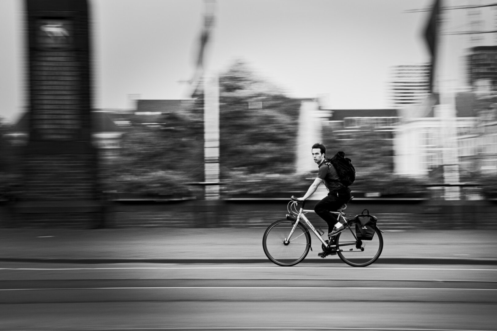 A man on a sportbike going full speed looks into the camera with an air of confussion. He's wearing fancy, shiny shoes and the rest of the outfit is sporty. He's also wearing a backpack. In the background you can see the blurred lines of the street a flag pole and a stone pillar. Further back you can see the faint outlines of the city skyline. Shot in black and white, tracked to the biker.