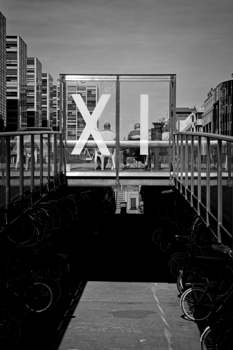 Black and white photograph of the arch running over the bike stall at Leiden Central Station. Two gaurdrails span over the dark bike stall leading to the center with a plexiglass wall with the letters X I. Behind the wall three men sit with their backs to the camera, enjoying the sun.
In the foreground a bright squar of sunlights fall into the bike stall below.