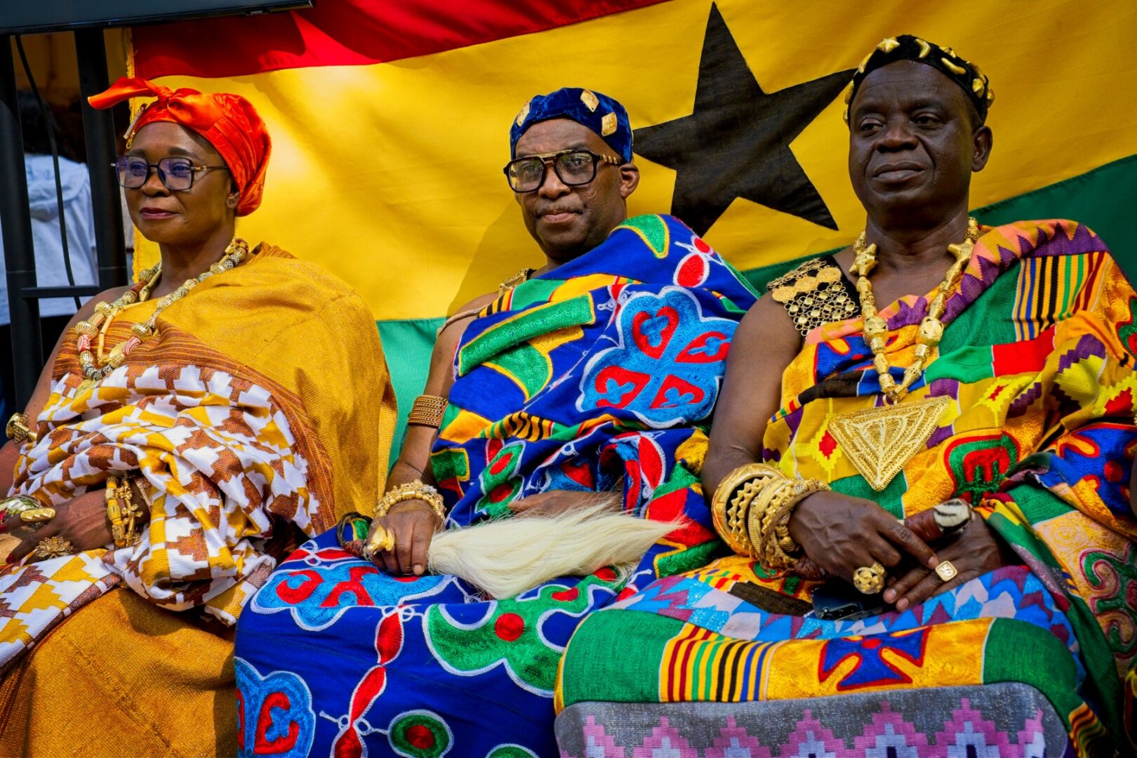 An african women and two men sitting infront of the Ghana flag in bright colored cloth wraps wearing gold jewelery. The scene has a regal atmosphere.