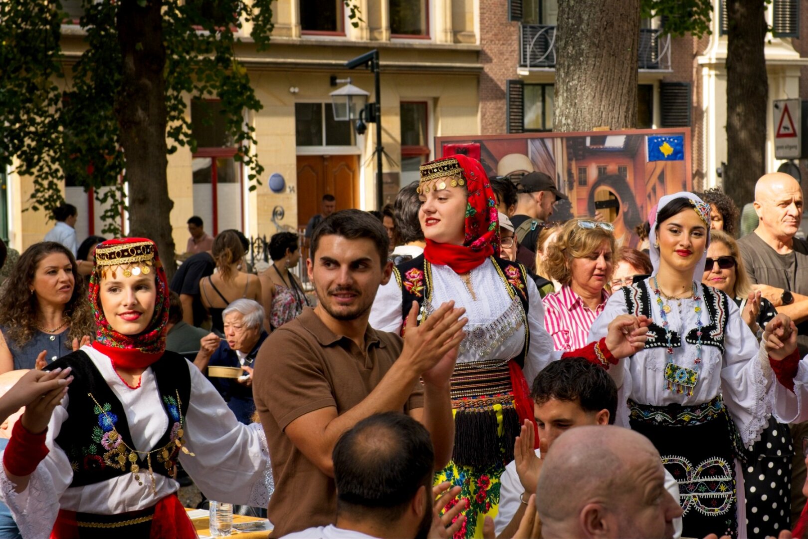 A busy street packed with people where three woman are forming a circle with the crowd to dance. Two of them are wearing red flowery scarves around their heads with gold embroidery and the one on the right is wearing a simpler white scarf.
All of them have black coats with bright white puffy shirts embroidered with roses and asorted flowers. The woman in the middle is standing tall amongst the crowd of clapping people.