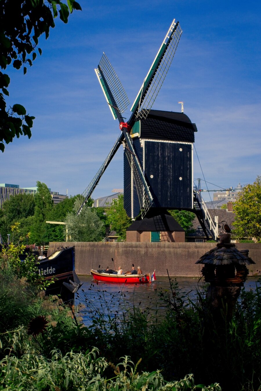 De Put windmill (wood, green, squar shaped), on a bright sunny day. 
A tree and some shrubs in the foreground, along with a birdhouse with a bird icon on top. A bright red boat floats past in the canal infront of the windmill