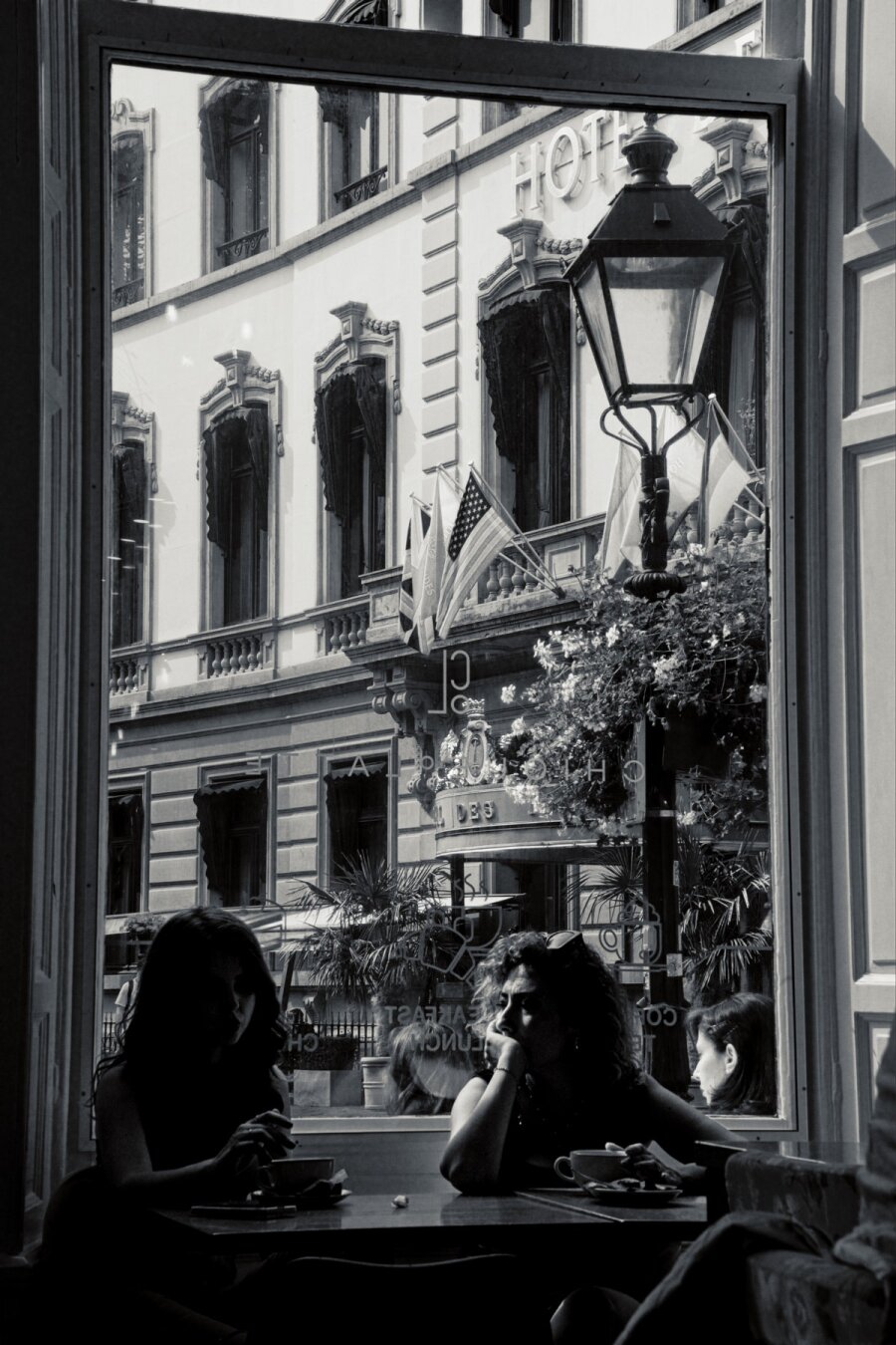 Black and white photo of a luxery hotel (build in 1858 as a palace) seen through a window. At the bottom of the window are two silloutes of women having a drink behind a table.
In the right side of the window an old timy street latern is visibile.