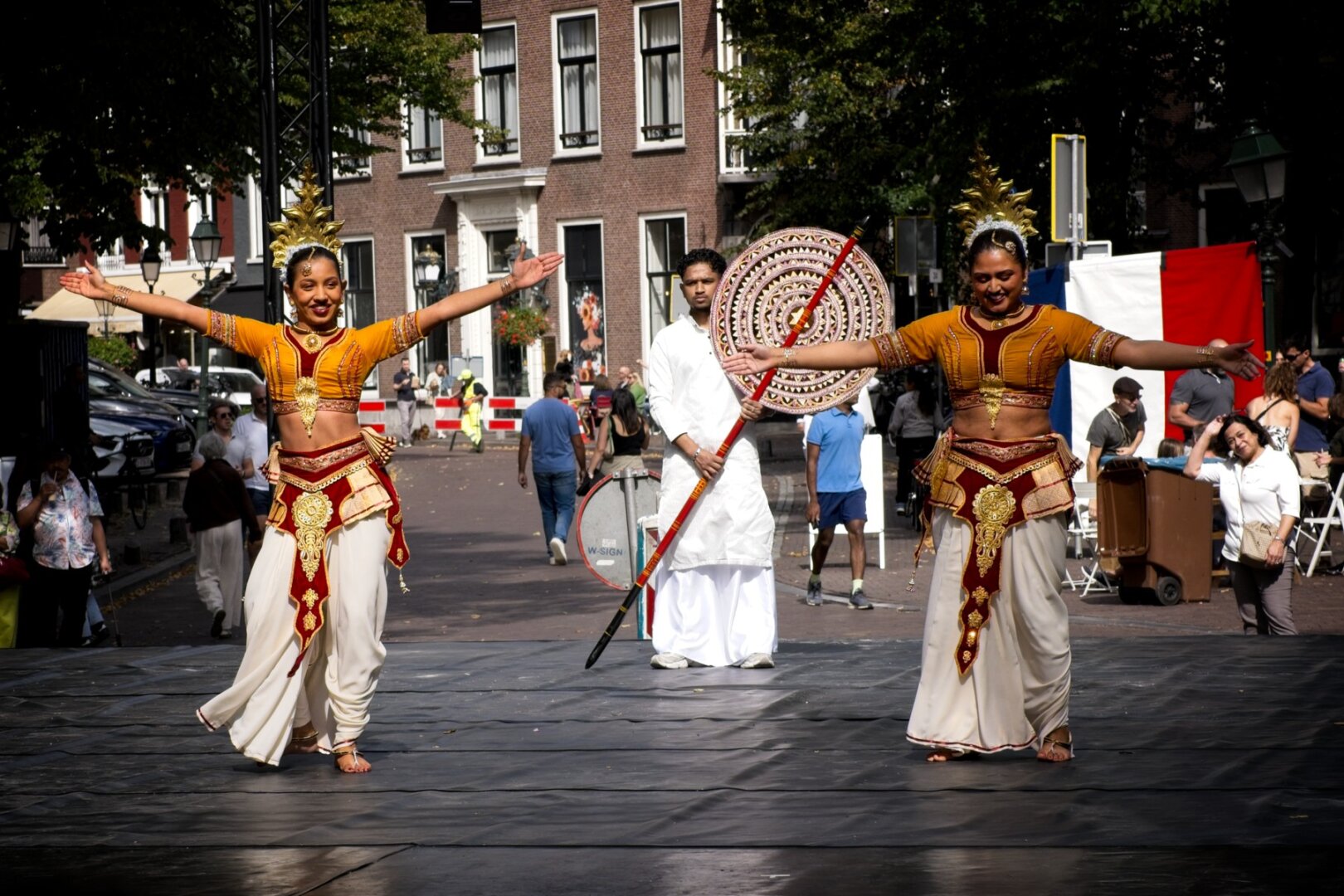 Two sri lankan dancers wearing gold headsets and white baggy skirts standing on a black stage with their arms spread, mid dance move. A man holding a staff with a woven shield stands behind them looking stoic. In the background is a street where bystanders are walking along.