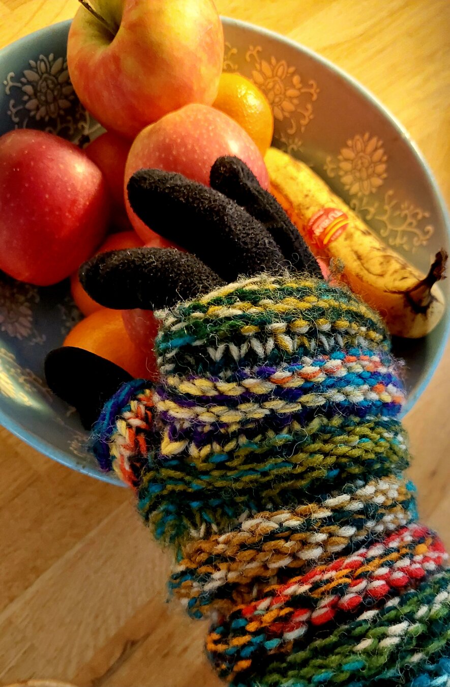 A black glove with a thick knitted fingerless glove over it in beautiful warm colors red orange yellow blue green. The glove holds an apple from the light blue fruit bowl