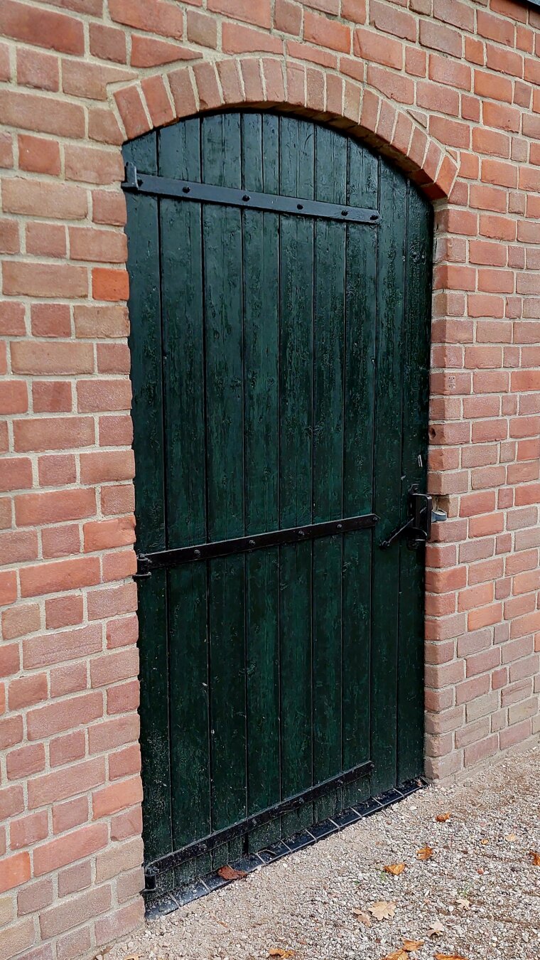 Dark green, old, wooden, neatly painted door, a wall.of bricks around it. The wall is cleaned too. All very neat, bright and shining.