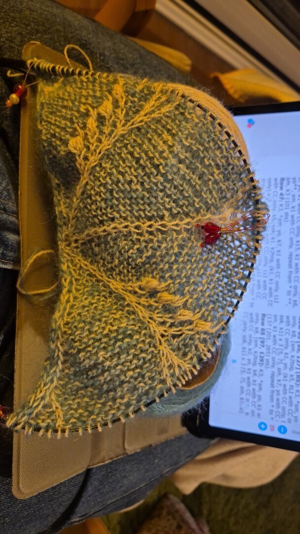 Ein Strickstück mit filigranem hellen Muster auf türkisblauem Untergrund liegt auf zwei Wollbobbeln in entsprechenden Farben, die auf der ausgeklappten Hülle eines Tablets stehen. Das Tablet mit der Strickanleitung ist dahinter aufgestellt.