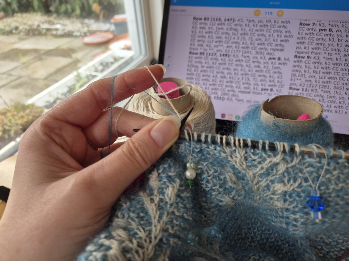 eine Hand mit zwei Fäden über den Zeigefinger halt ein Strickstück. im Hintergrund sind die beiden Garne auf Papprollen aufgewickelt und dahinter ein aufgestelltes Tablet mit der Anleitung eher rechts und links der Blick auf die Terrasse.