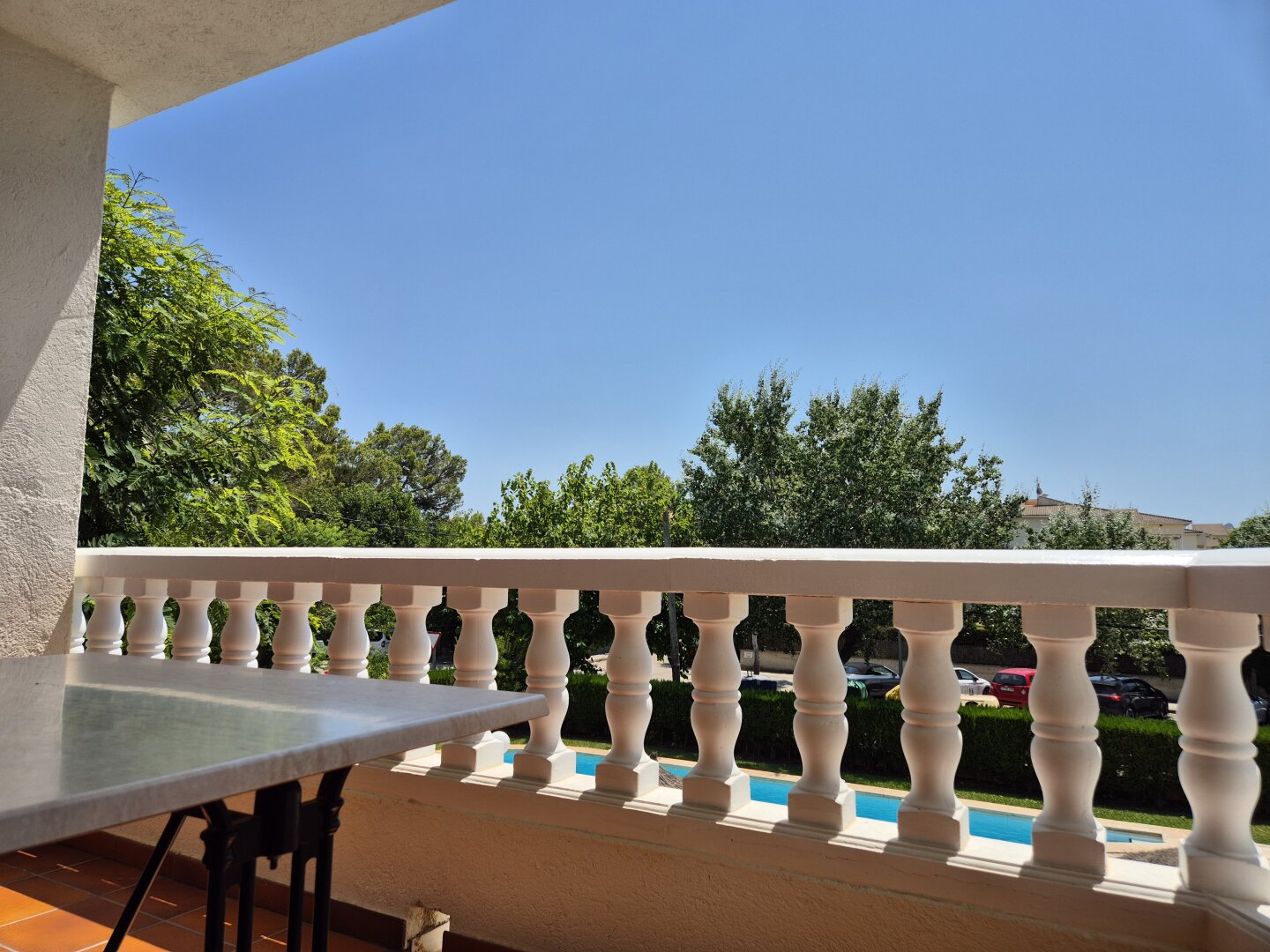 Blick von einem Balkon halbhoch in den Himmel. Man sieht locker im Bild angeschnitten einen Tisch, dahinter fast auf ganzer Breite eine durchbrochene Balkonbrüstung, durch die man einen Pool erahnen kann, dahinter Bäume und darüber klar blauen Himmel
