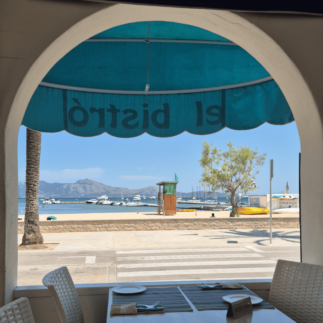 Blick von einem Restaurant nach draußen. Hinter der Strandpromenade ist das Meer, am Horizont ist eine Bergkette, darüber blauer Himmel