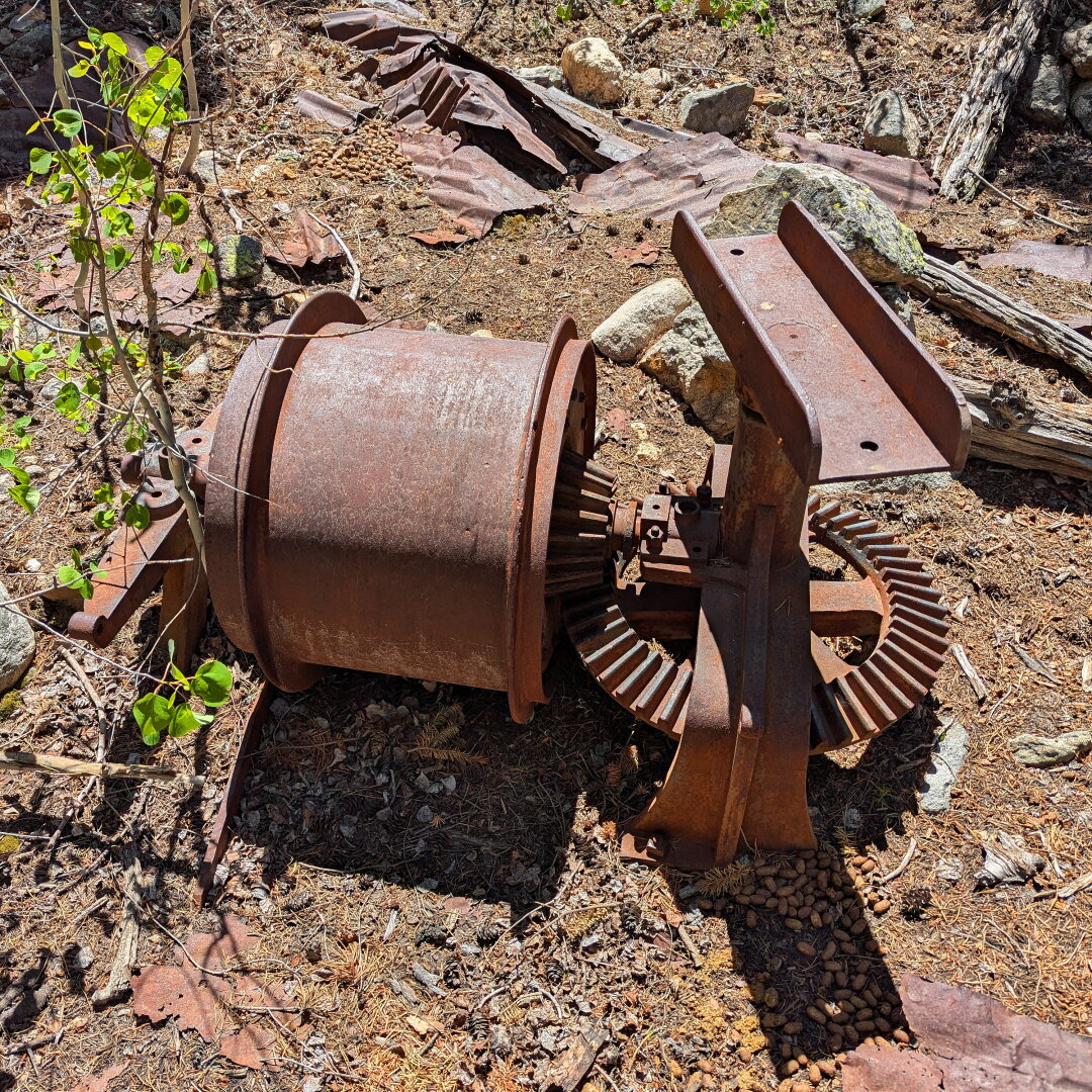 An old, rusted piece of machinery with a large cylindrical body and gears, lying on the ground surrounded by debris and plants.