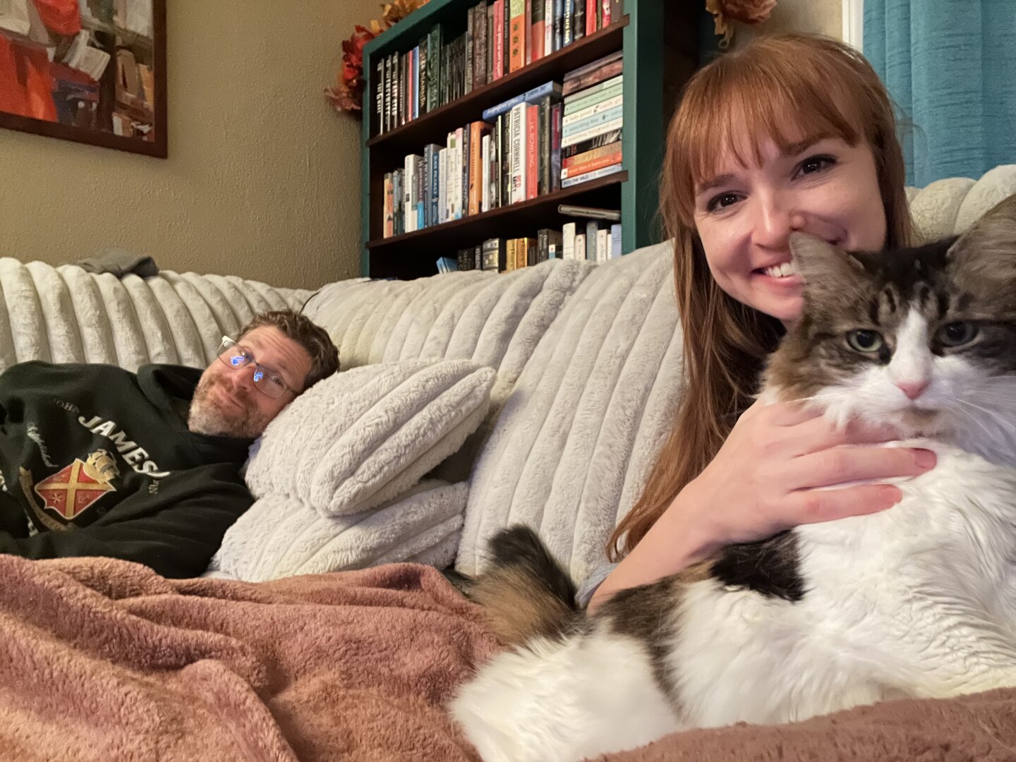 Human dad, human mom and a fluffy white and brindle cat pose for a family photo on a cozy cream couch.