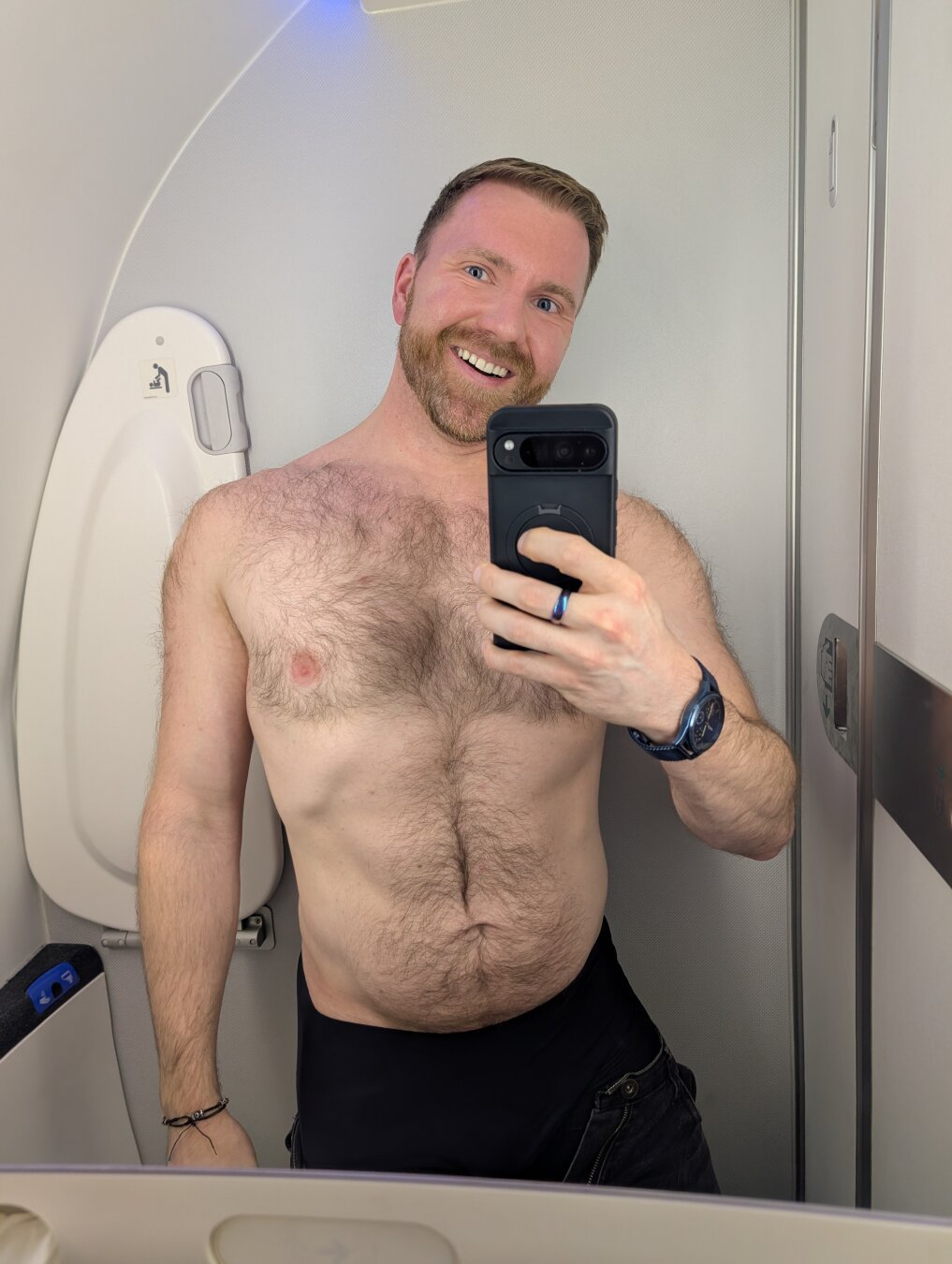 Smiling bearded man in an airplane bathroom, shirtless showing chest hair.