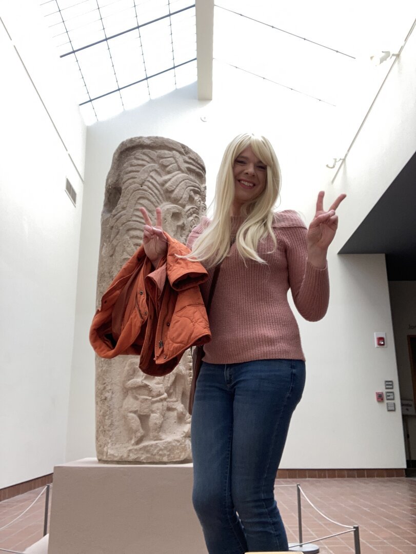 Blonde woman in pink sweater and jeans posing for a selfie in front of sculpture