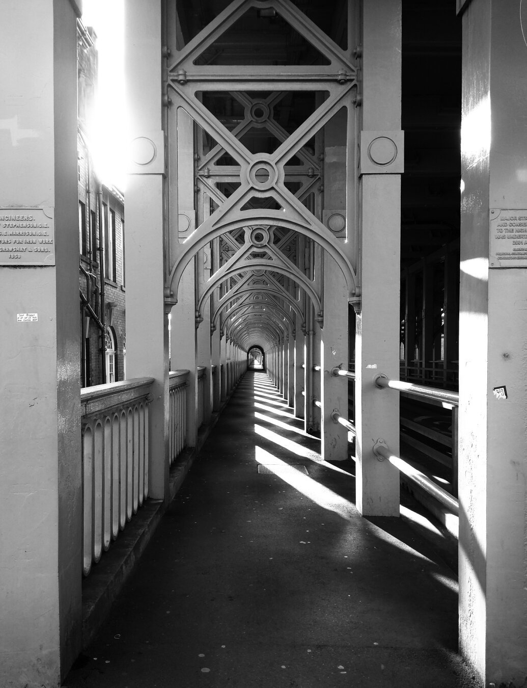 A black and white photo showing a walking path through a bridge. The bridge has a top level, so the path is covered. There are white spans holding up the deck receding into the centre of the photo. It's quite early in the morning, and the sun is coming in at an angle the shadow from the handrail covering half the path, the other half has black and white stripes from the spans