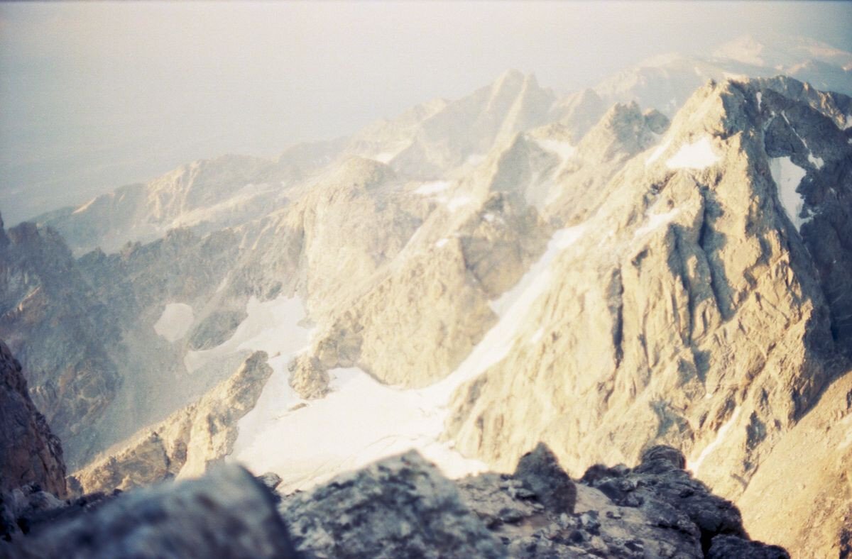 A panoramic view of rugged mountain peaks, specifically the Middle Teton, under soft, diffused light. The landscape features rocky terrain with patches of snow, showcasing the texture and contours of the Teton Range. The image has a slightly blurred or hazy appearance, emphasizing the grandeur of the mountains.