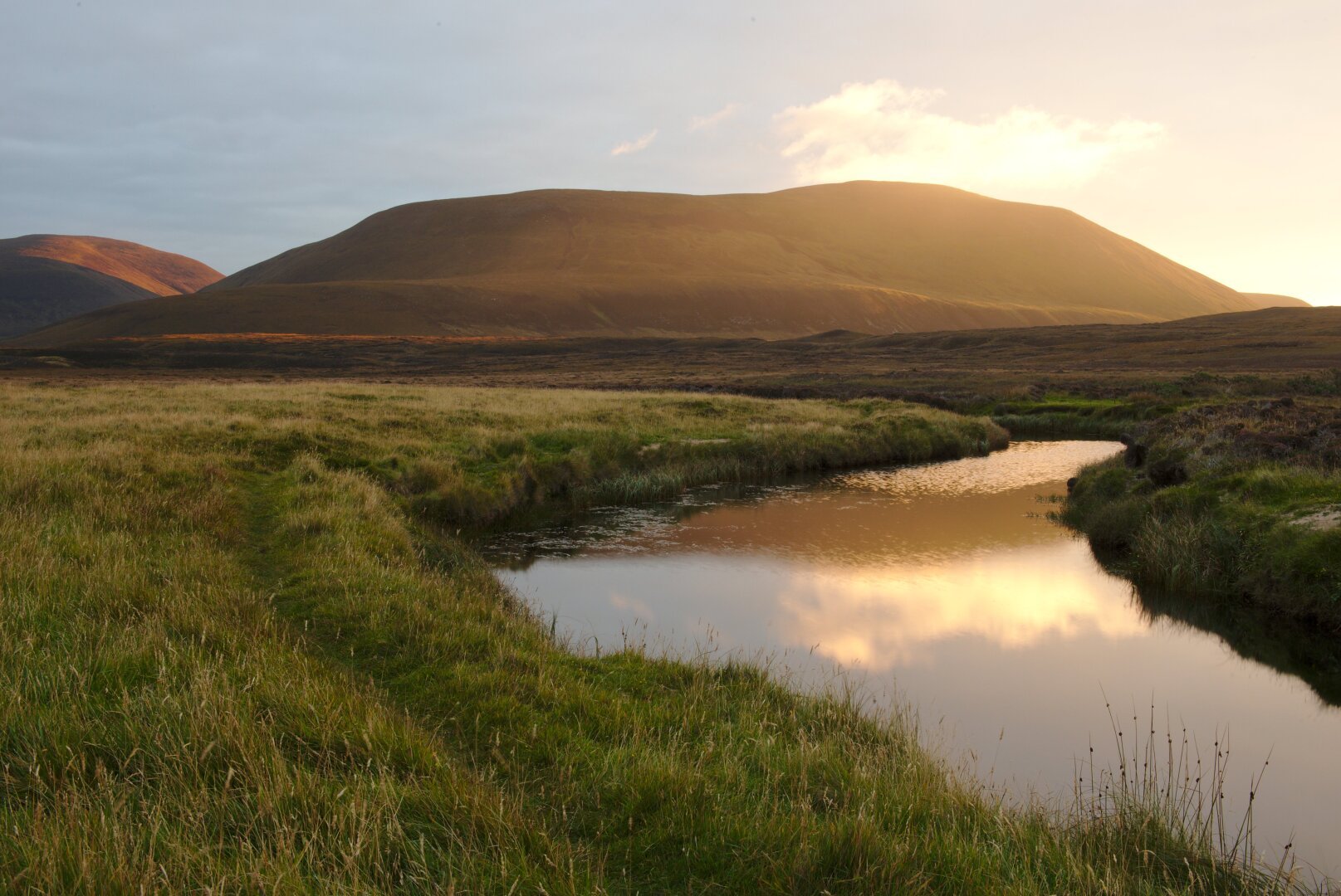 The image captures the Isle of Hoy at sunset, with the setting sun casting a warm golden glow over the rolling hills and grassy landscape. A gently winding stream reflects the soft hues of the evening sky, creating a mirror-like effect. The surrounding terrain, covered in heather and grass, is bathed in the fading light, emphasizing the contours of the land. The scene exudes a sense of tranquility and untouched beauty, characteristic of Hoy's rugged and remote wilderness.