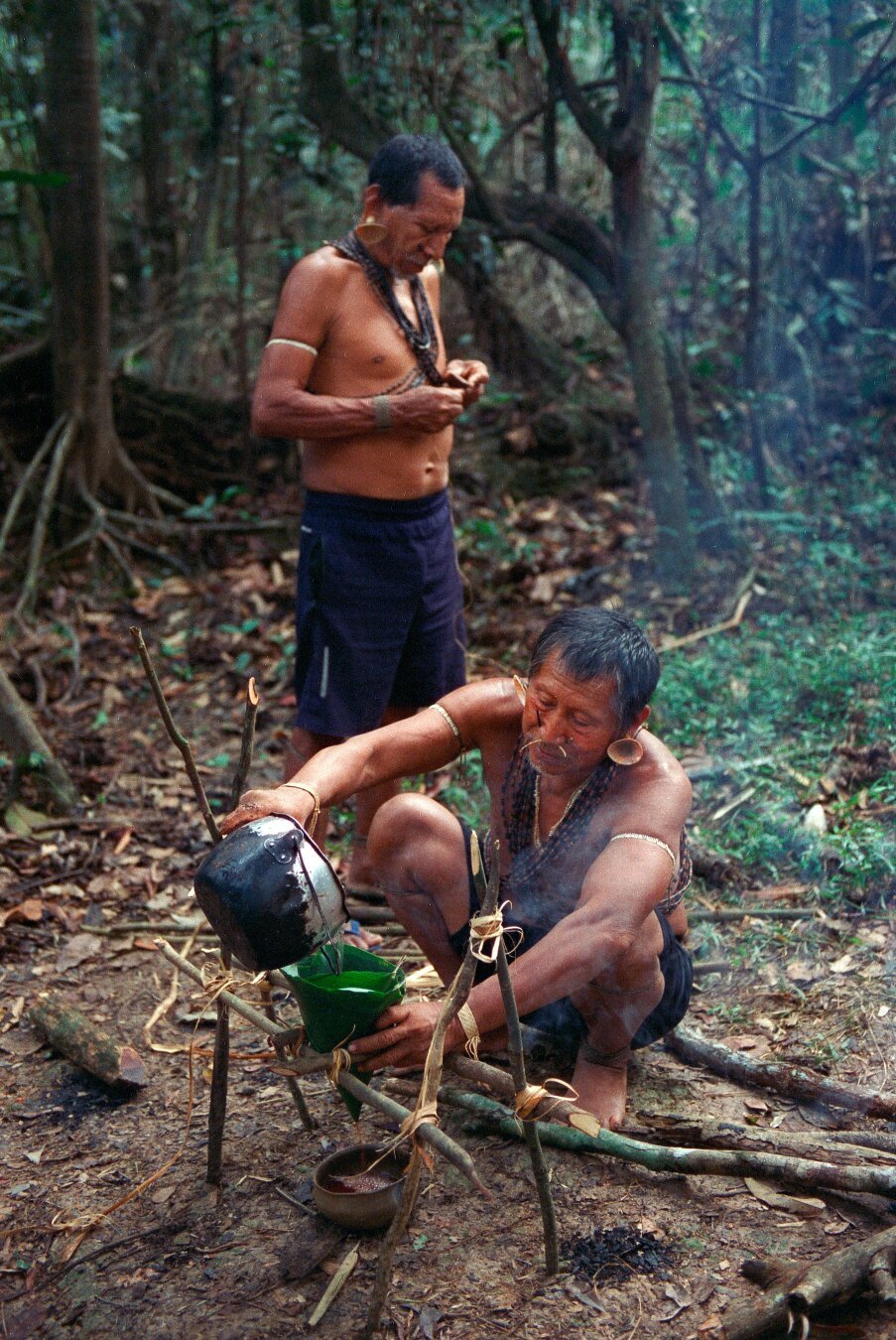 Two indigenous Matis men in a dense rainforest prepare curare, a traditional poison used for hunting. One man, wearing body adornments including necklaces and arm bands, is squatting near a small fire, carefully pouring liquid from a blackened pot into a green leaf. The other man, standing behind him, is focused on crafting or preparing an item in his hands. The surrounding forest is lush with trees and foliage, creating a natural setting for their work.