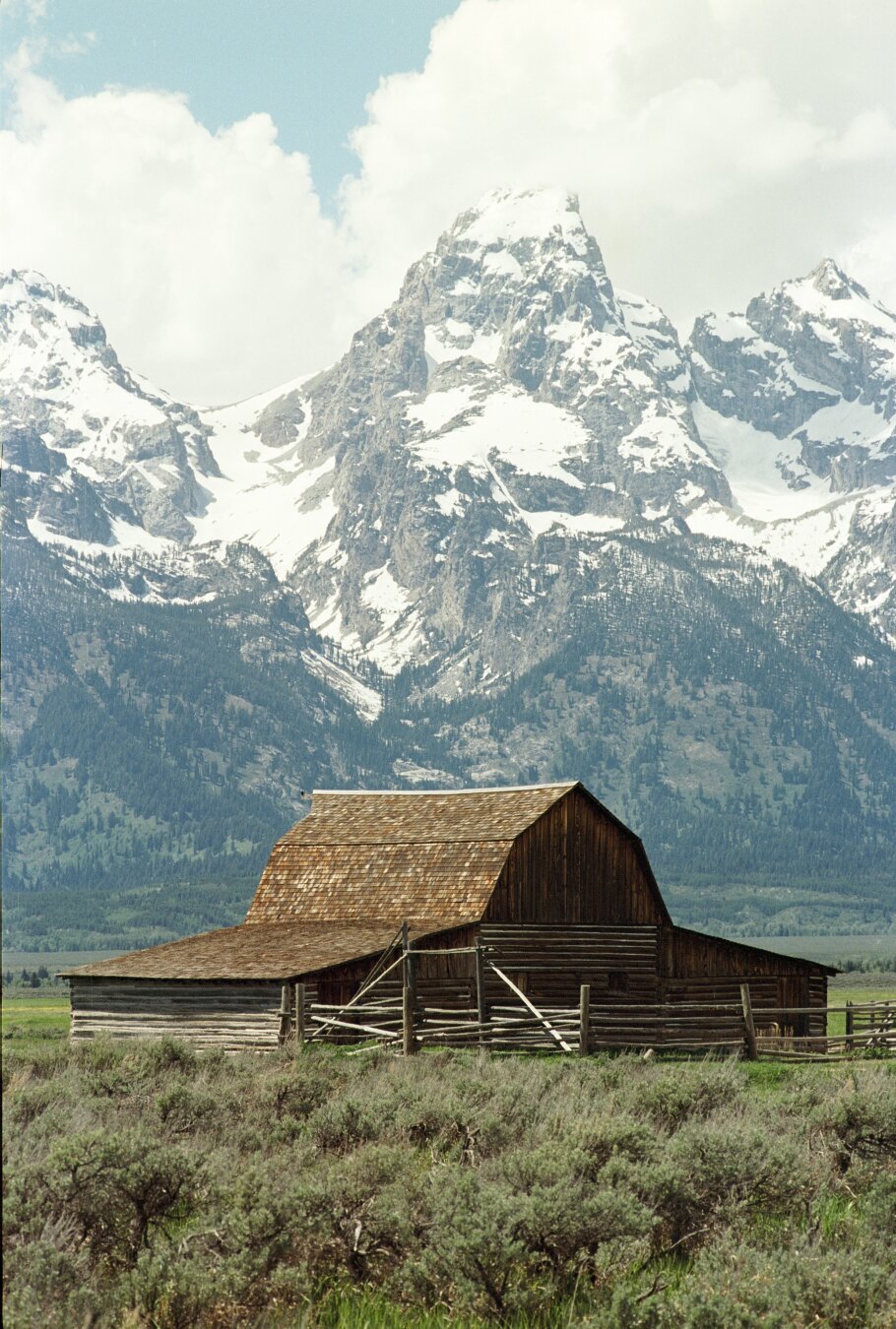 A historic wooden barn with a shingled roof stands in an open field, enclosed by a weathered wooden fence. In the background, the majestic, snow-capped peaks of the Teton Range rise sharply against a partly cloudy sky. The foreground is dotted with sagebrush, adding to the rustic, picturesque scene of Mormon Row in Grand Teton National Park.