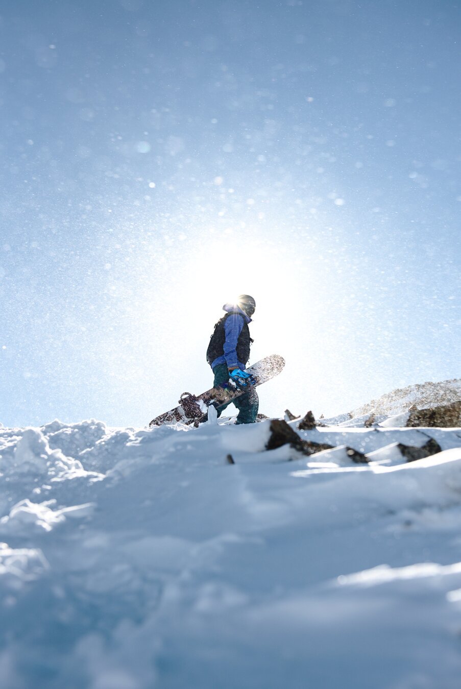 A snowboarder dressed in winter gear is hiking up a snow-covered mountain with a snowboard in hand. The sun shines brightly behind them, creating a backlit silhouette effect, while snowflakes scatter in the air. The terrain is a mix of snow and exposed rocks, indicating a rugged alpine environment.