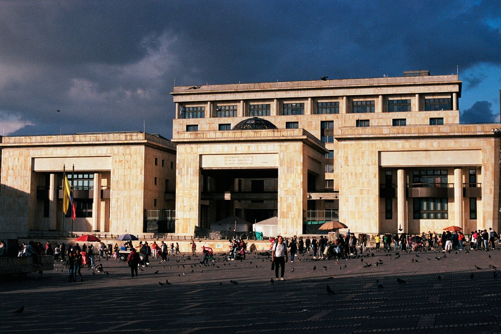 This image shows a large government building in Plaza de Bolívar, Bogotá, Colombia. The structure features a modern neoclassical design with large rectangular columns and beige stone facades. A Colombian flag is visible on the left side, and many people are walking around the plaza, with pigeons scattered across the open space. The sky has a mix of blue and dark clouds, casting dramatic lighting on the scene.