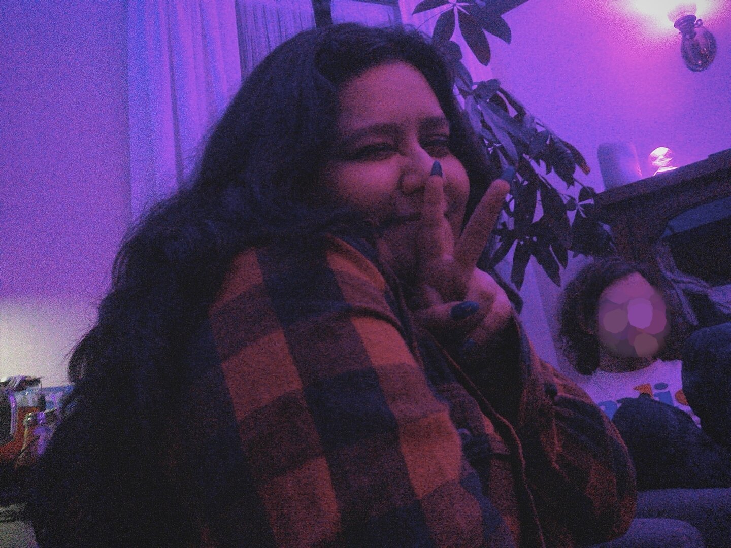 one sexy bish she they peace sign at a party with long black wavy hair work winking at camera chola sweater purple lighting plant behind her blurred face in background