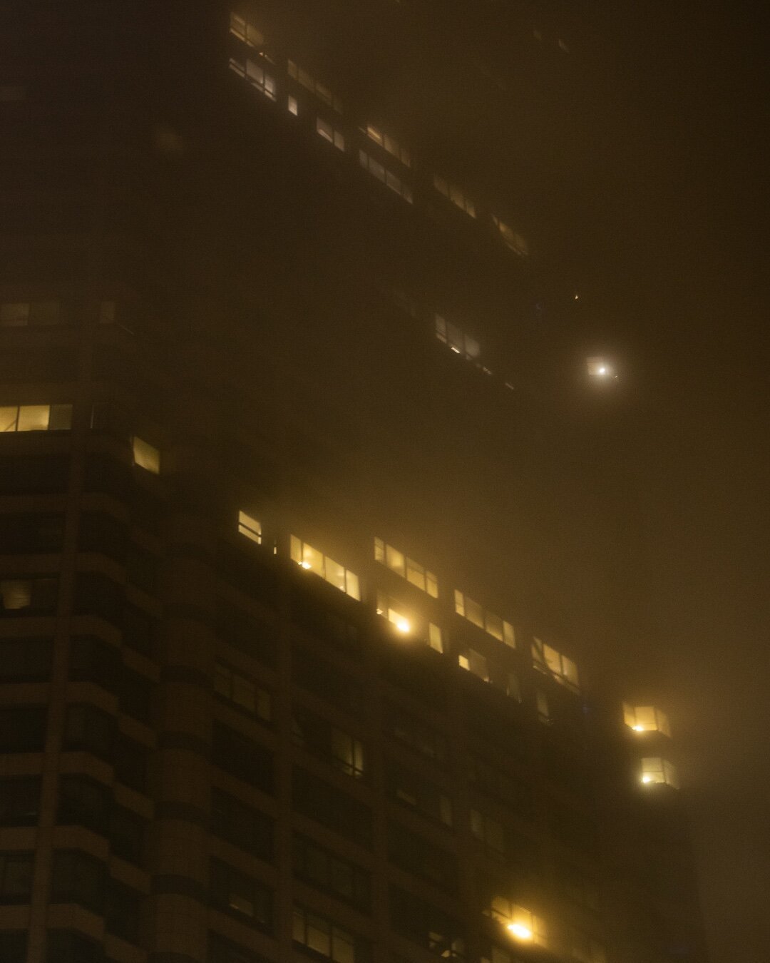 The Seattle Municipal Tower at night. Captured from below, the surrounding fog scatters the light from each window, spilling out the light into a glow around the building.