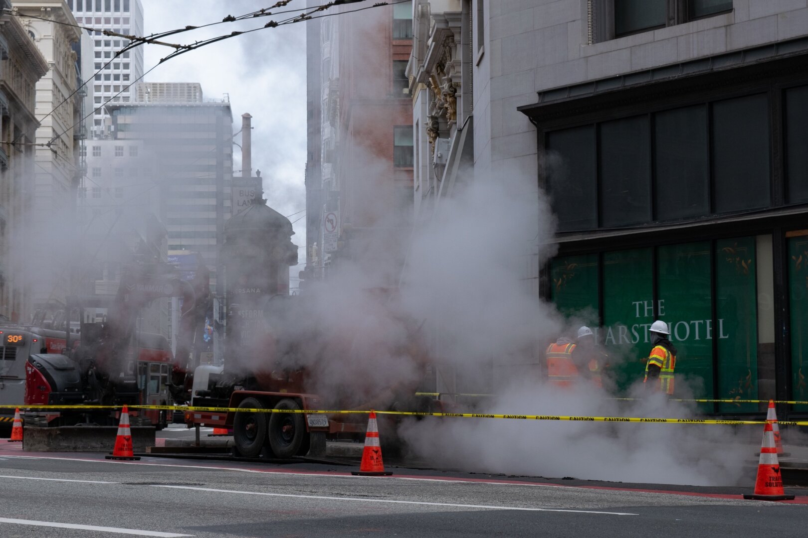 Landscape image of street construction in San Francisco. Steam emits from some exposed underground piping, just out of view in below right frame. Three men in orange-and-yellow construction vests, with two also covered by the steam, are in discussion, presumably about the construction (though really could be about anything).